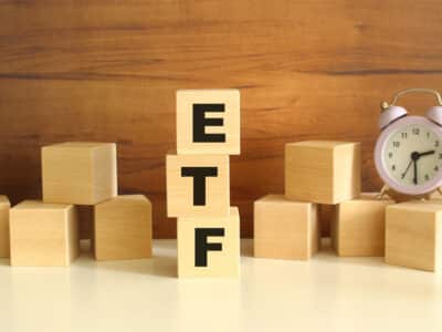 Three wooden cubes stacked vertically on a brown background make up the word ETF. Cubes are scattered nearby and there is a clock. Front View Concepts